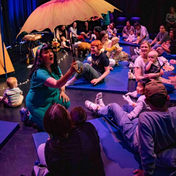 A female performer kneels on the ground amidst an audience of babies and their parents all sat on the floor in a theatre. The performer is singing and holding a flower-shaped umbrella.