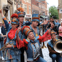 Five performers dressed in red and blue marching band outfits. They are standing on the street surrounded by crowds. They are making silly faces and some are holding brass instruments.
