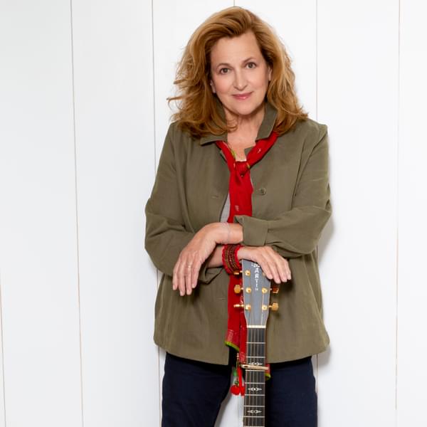 Singer Barbara Dickson is stood against a white wall leaning on a guitar. She is Caucasian, has shoulder length blonde hair and brown eyes. She is wearing a green blouse, red scarf and black trousers.