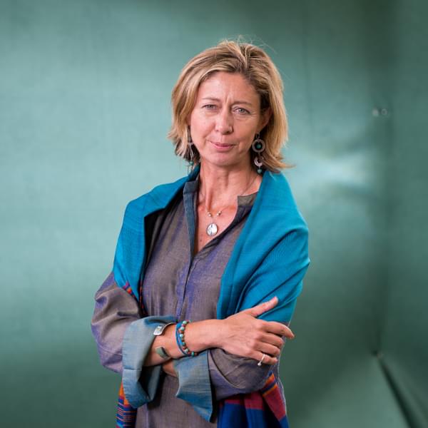 Journalist Christina Lamb is standing with her arms crossed against a teal blue background. She has short blonde hair, blue eyes and is Caucasian. She is wearing a grey blouse, blue shawl and chunky jewellery.