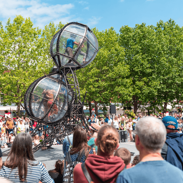 Four circus performers in a large hourglass apparatus. They are performing in Market Square in Salisbury and are surrounded by crowds. There are trees in the background and blue sky.