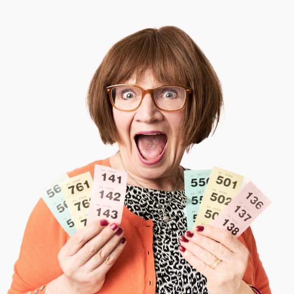 Janice Connolly as her character Barbara Nice. A middle-aged Caucasian woman wearing glasses and an orange cardigan. She is holding up 6 strips of raffle tickets.,