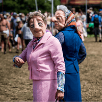 Two performers wearing eccentric and exaggerated elderly lady masks, wigs and twin-set suits walk through a music festival. They are outside in the sun, one is looking into the camerea, another is facing away from the camera, showing their side profile.