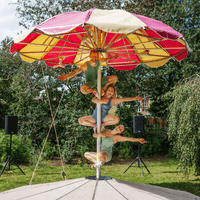 Three circus performers dressed in green one-piece swimming costumes are hanging on to the pole of a giant beach parasol. This parasol is red and yellow and is outside in the grass on a sunny day.