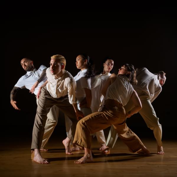 Six dancers is white button up shirts strike various poses. The background is black and they are standing on a wooden floor.