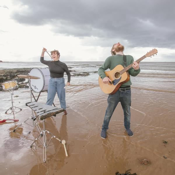 Two musicians, one female and one male, stand playing their instruments in the shallow water on a beach. They are screaming at the sky. They are drums and small instruments in the sand. They are wearing casual clothing.