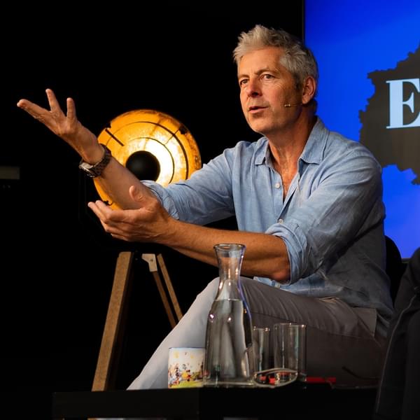 Author Justin Webb sits on a chair on stage giving a talk. He is wearing a blue shirt and is gesturing with his arms while talking. He is Caucasian with brown eyes and grey hair.
