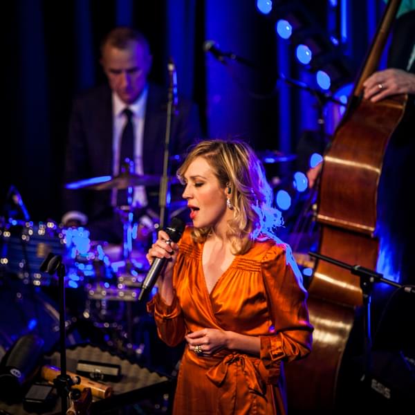 Lucy Randell wearing an orange dress on stage singing into a microphone. There is a drummer and double bass player slightly blurred in the background