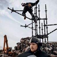 Three dancers dressed in black athletic jumpsuits and leather caps stand in a junkyard with a bulldozer in the background. Two of the dancers are blurred in the foreground in an embrace, the other is striking an acrobatic pose on a metal frame that forms the basis of their show.