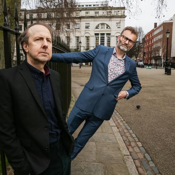Steve Punt and Hugh Dennis on a pavement next to some railings. Punt is wearing a dark suit with his hands in his pockets and Dennis is wearing a light blue suit and is holding onto the railings leaning at an angle