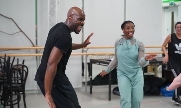 Actors Jammy Kasongo and Chioma Uma are both smiling and dancing during rehearsals for ‘Brief Encounter’. Jammy is wearing a black t-shirt and black jogging bottoms, and Chioma is wearing light blue dungarees.