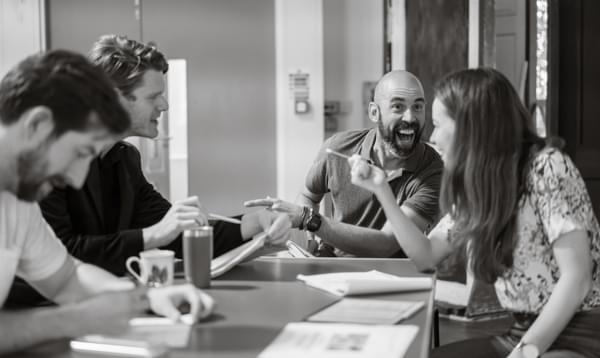 A black and white shot of actors Patrick Warner, Luke Barton and Alistair Cope with director Marieke Audsley, all smiling sitting round a table reading scripts. Alistair is looking at Marieke and laughing.