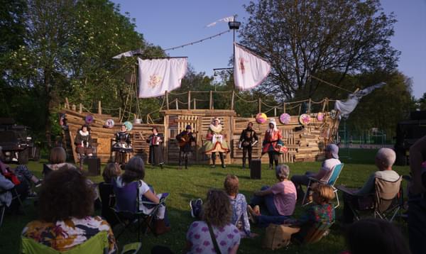 In front of a dusky blue sky and trees, a huge wooden ship made from recycled wooden pallets with seven actors in Shakespearean dress in front. The foreground has audience members sitting on the grass and in chairs watching the performance.,