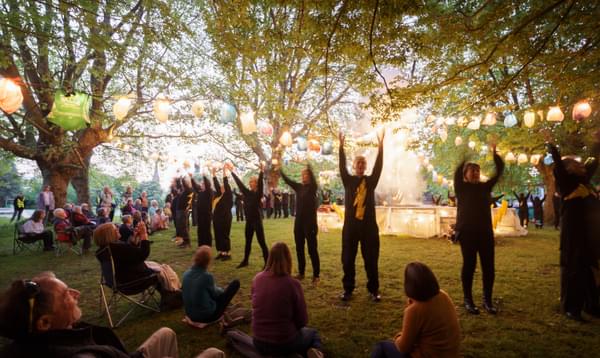 Community actors dressed in black with yellow lighting bolts on their front, stand in a line with their hands in the air. Behind them is a stage covered in lights, with festoon lanterns made from carrier bags hanging across to trees. Audience members are sat on the grass and chairs watching the performance